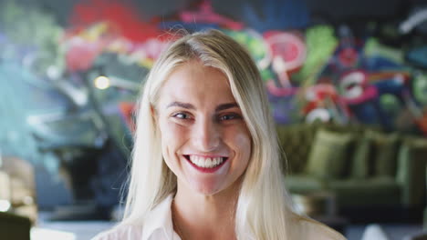 Young-white-woman-with-blonde-hair-in-front-of-mural-smiling-to-camera-and-laughing,-close-up