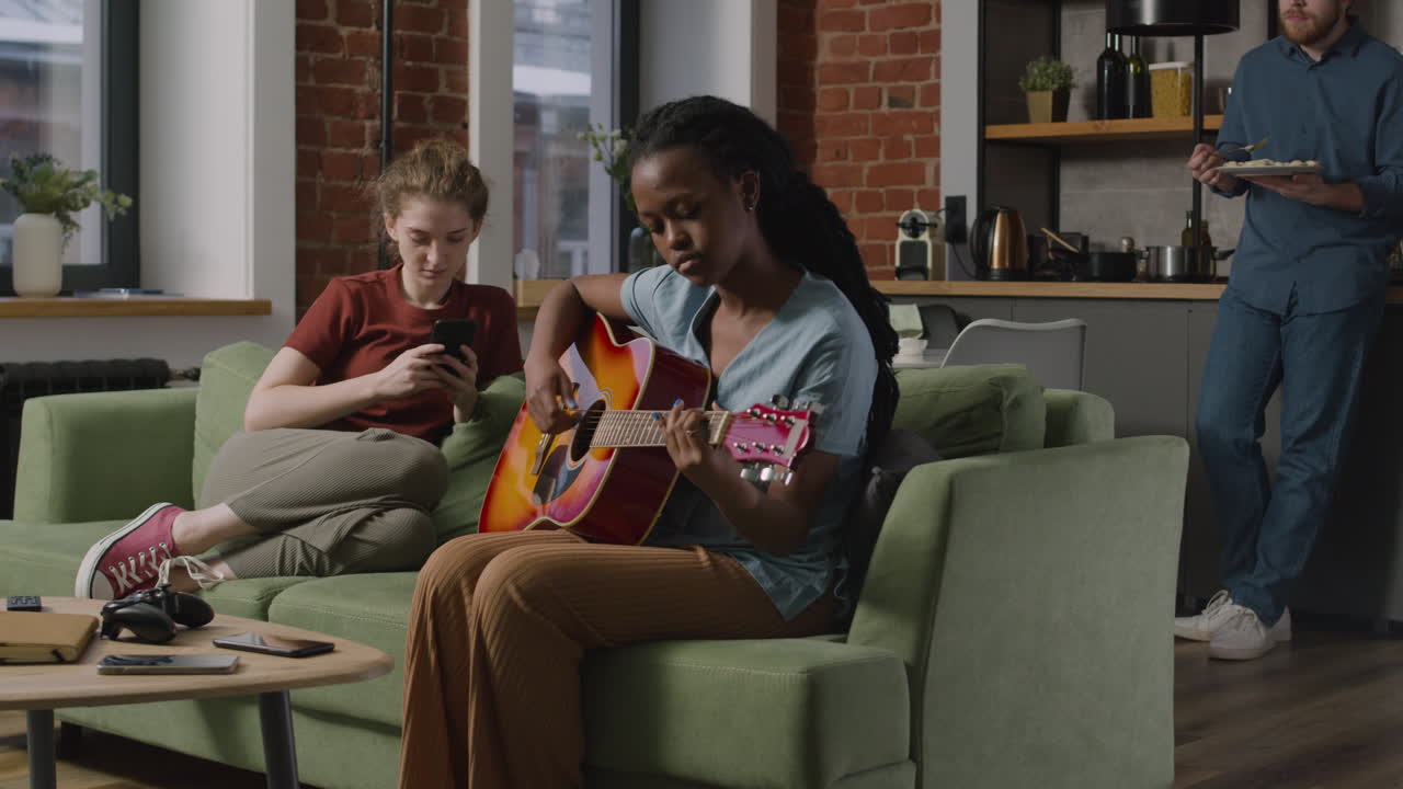 niña tocando la guitarra sentada en el sofá mientras su compañera de cuarto usa el teléfono inteligente