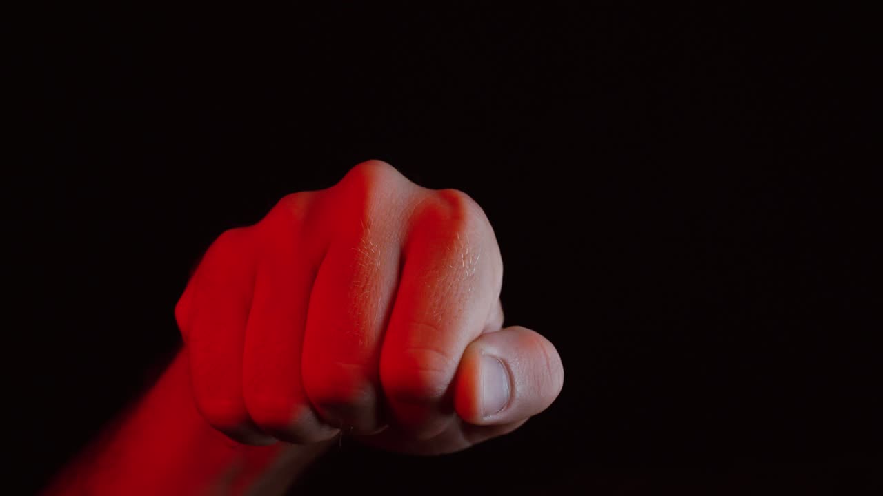 A close-up of a hand forming a fist and striking toward the camera under dramatic red lighting, symbolizing strength, anger, or confrontation in a cinematic dark setting