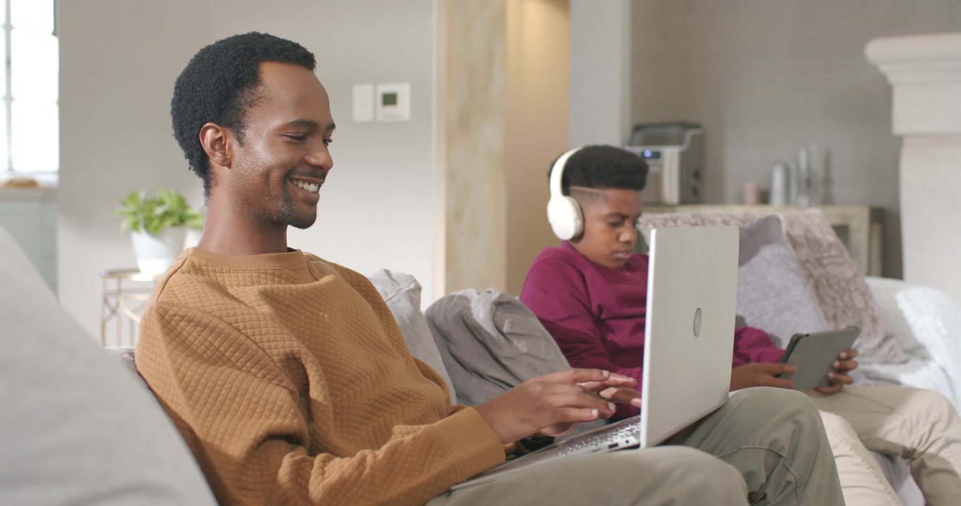 Father and teen son on couch, father using laptop while son tapping tablet wearing white headphones