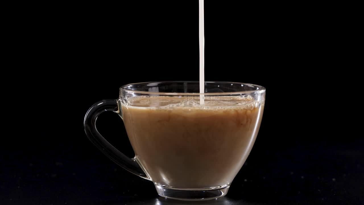 Milk streams into a glass cup of coffee, creating swirling patterns against a dark backdrop