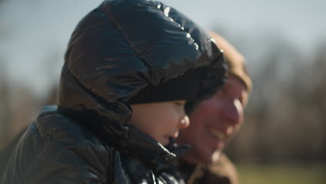 a close-up of a father and his son sharing a joyful moment together outdoors, the child, dressed in a shiny black jacket, laughs alongside the father, who is wearing a brown jacket