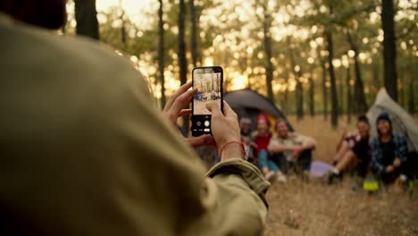 A-man-in-a-light-green-shirt-takes-pictures-of-his-company-with-whom-he-went-hiking-using-a-camera-on-his-phone-in-a-sunny-green-summer-forest.-Taking-a-photo-for-memory-during-a-camping-stop