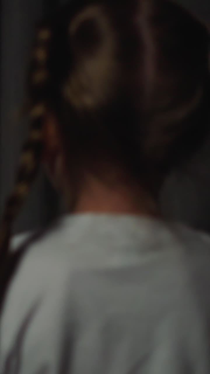 Blonde little girl with plaits worried of blinking flashlights looks out of window on street from dark room in evening backside view slow motion