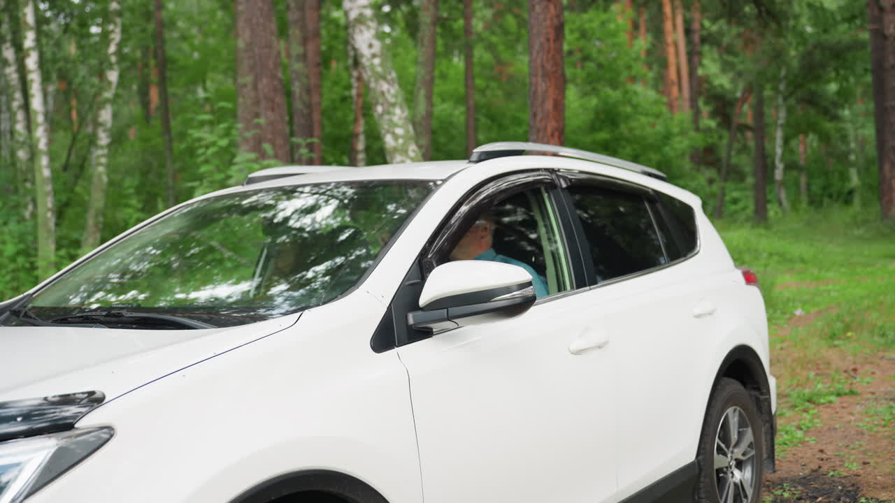 Two parents sit inside white SUV parked in lush green forest, surrounded by trees and calm summer atmosphere, reflection of nature visible on car glass, outdoor lifestyle moment