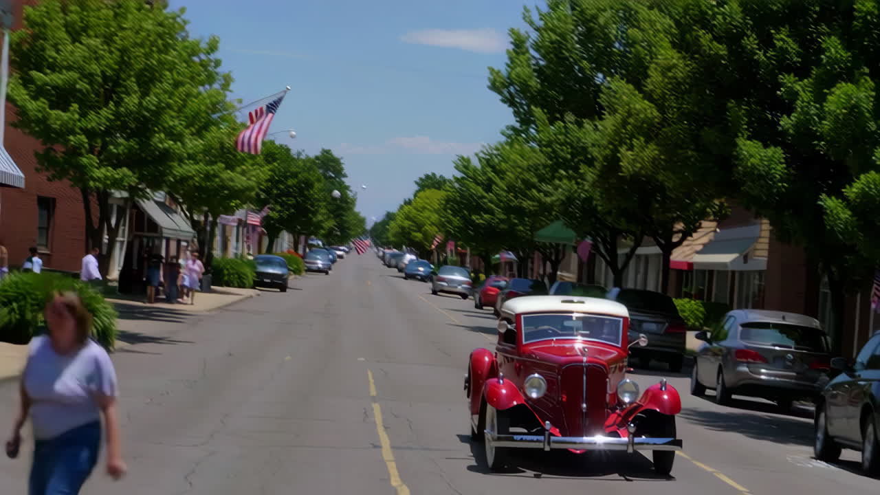 Vintage Red Car Driving Down a Main Street with American Flags and Trees on a Sunny Day