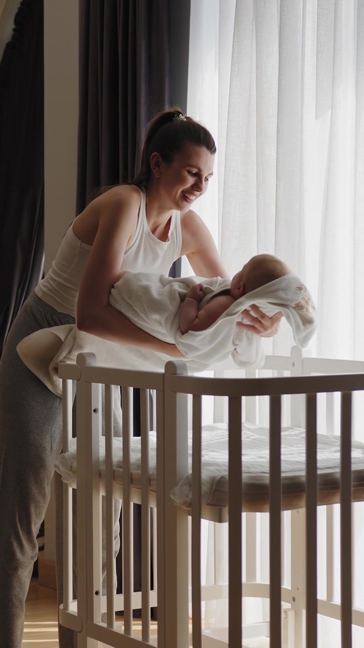 Young mother gently places swaddled infant into white crib near window with soft light filtering