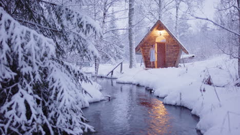 woman exit wooden sauna in towel near river in snowy woods as calming snow falls