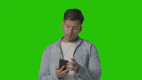 foto de estudio de un joven vestido informalmente enviando mensajes por teléfono móvil contra una pantalla verde