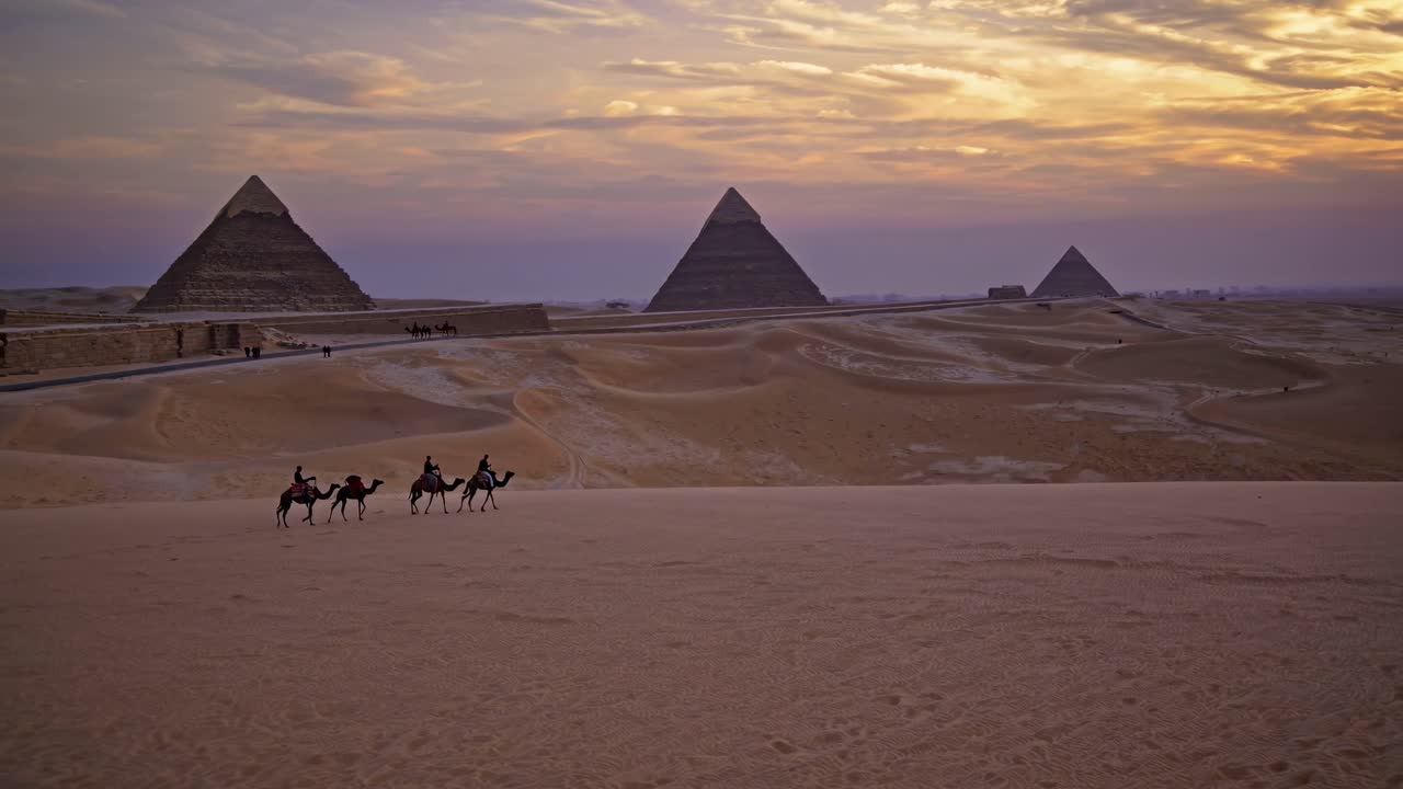 Wide-angle video shot of camels traversing the desert at sunset, with the pyramids