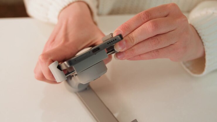 Close-up view of hands adjusting caliper jaws around small metal hinge while steadying component on smooth white tabletop, showing active precision measurement, alignment, and workshop focus