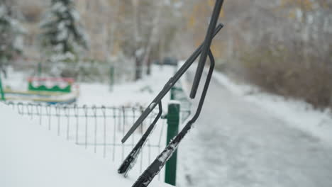 close-up of icy windshield wipers on a snow-covered car near a green metal fence in a snowy park setting, background features dry trees and blurred car moving in the distant