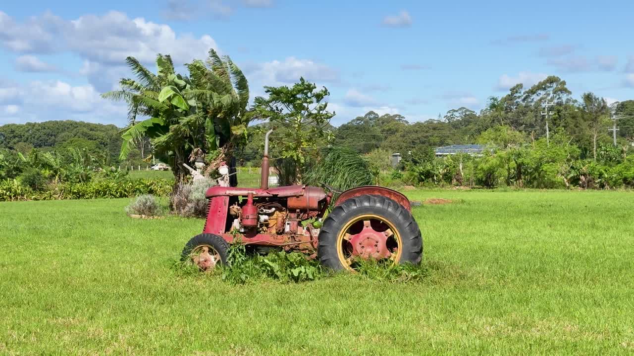 A stationary, weathered red tractor sits in a lush grassy field under bright daylight, with slow lateral camera movement revealing rural Byron Bay scenery