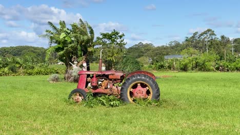 a stationary, weathered red tractor sits in a lush grassy field under bright daylight, with slow lateral camera movement revealing rural byron bay scenery