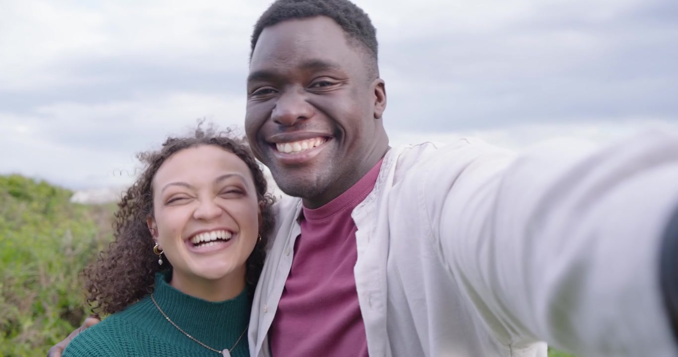 Casal afro-americano entrando no enquadramento, homem levantando a câmera, filmando um 'olá' nas dunas, brincos de argola