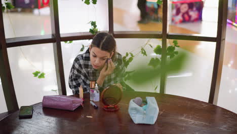hermosa chica aplicando maquillaje en su cara mientras usa un espejo, rodeada de artículos cosméticos y teléfonos inteligentes cuidadosamente dispuestos en una mesa de madera, vides verdes enmarcan un entorno pacífico en un espacio interior moderno