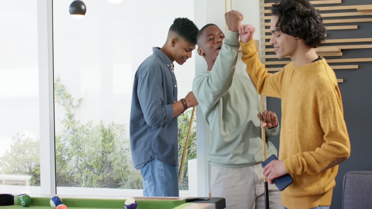 African American man celebrating shot by dancing with pool cue at table, friends cheering in lounge