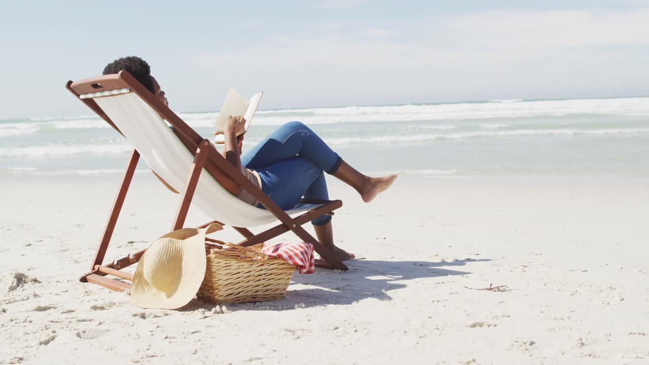 mujer afroamericana leyendo y acostada en una cama de sol en una playa soleada