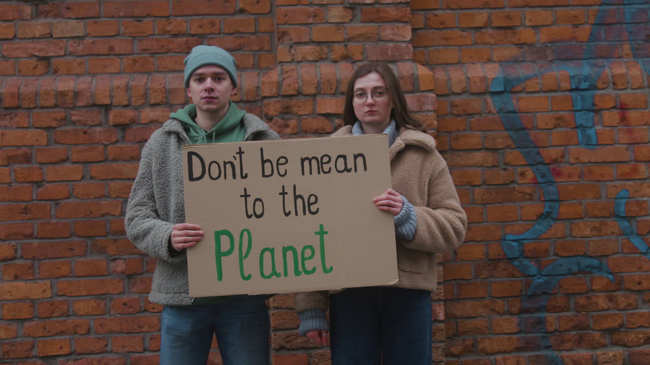 молодые мужчина и женщина активисты смотрят в камеру, держа картонный плакат во время акции протеста против изменения климата