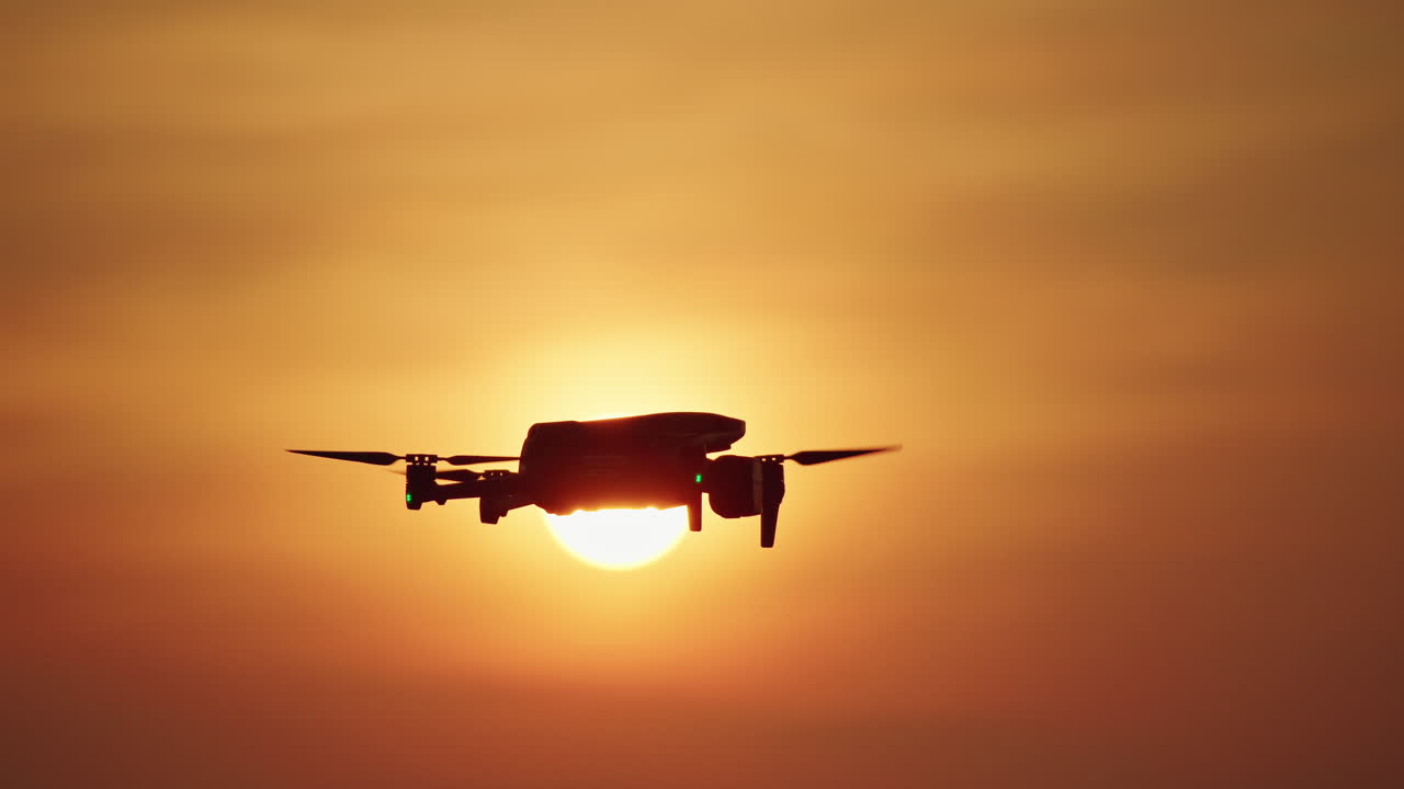Silhouette di drone che vola davanti al sole splendente al tramonto con cielo arancione.
