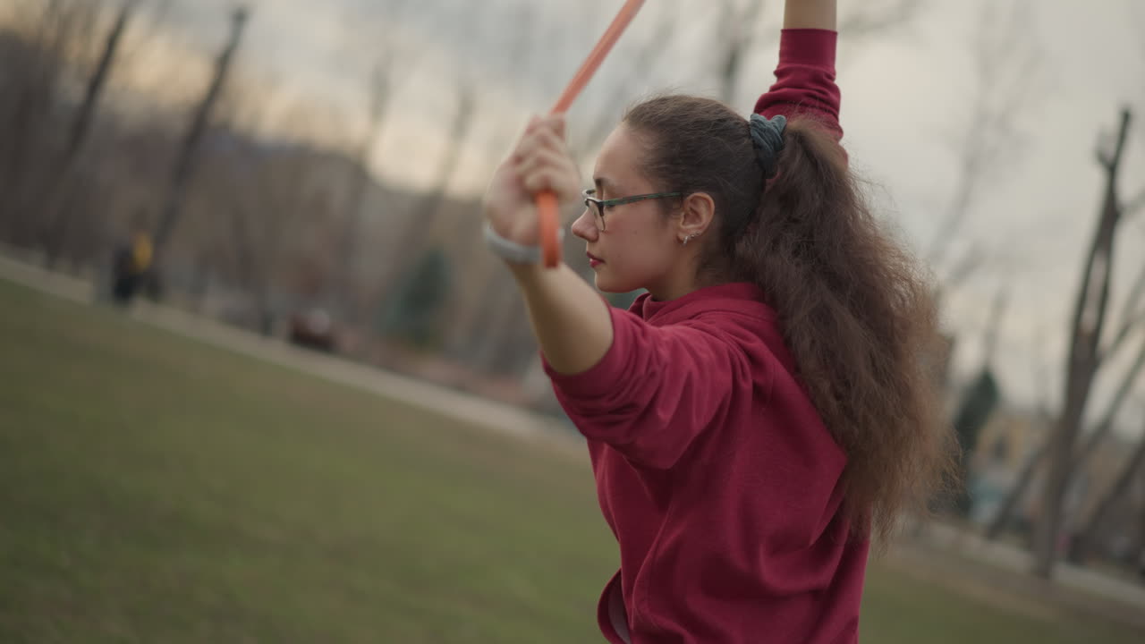 Young White Woman Stretching With Band In Park, Student Performing Slow Mobility Routine For Shoulder And Chest Using Orange Resistance Loop, Cloudy Light, Casual Hoodie, Glasses, Grass Field And Calm