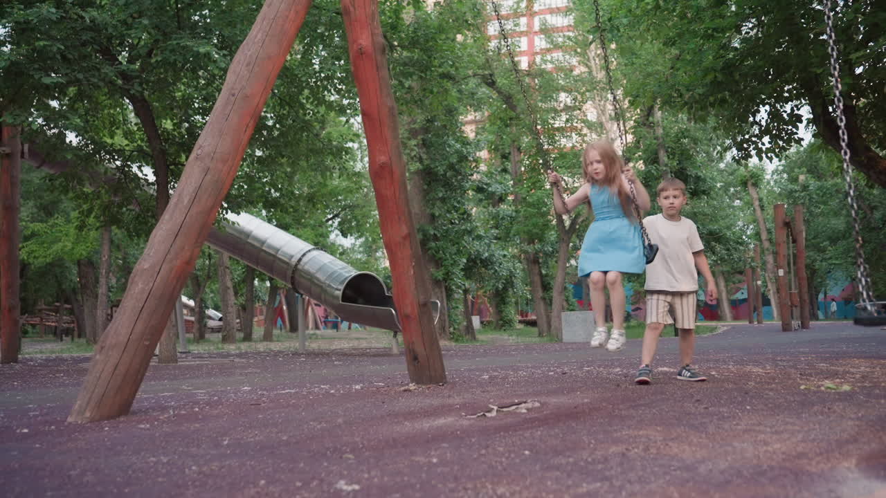 teenage girl smiles as she swings on wooden chain swing while playmate pushes from behind in lush park at dusk capturing joyful sibling play under warm evening sunlight shimmer