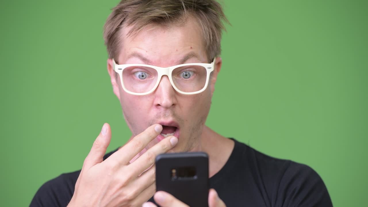 Young handsome Scandinavian nerd man looking shocked while using phone