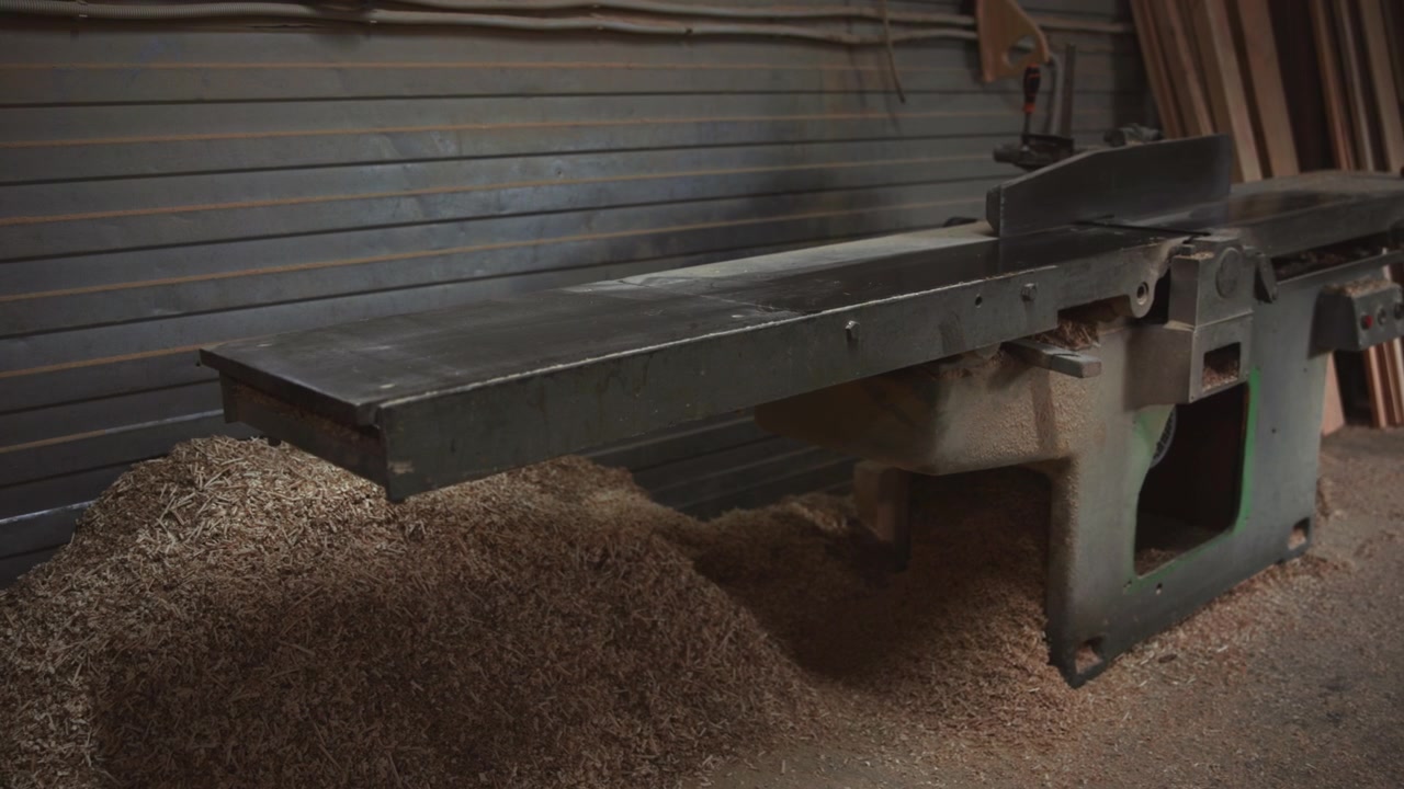 Low-angle panoramic view of long jointer planer inside woodworking shop, showing worn infeed surface, raised fence, chipped cast-iron body, scattered wood shavings, and textured floor