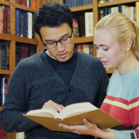 Hombre-Asiático-Con-Gafas-Y-Mujeres-Caucásicas-De-Pie-Juntos-Mirando-El-Libro-En-La-Biblioteca-1