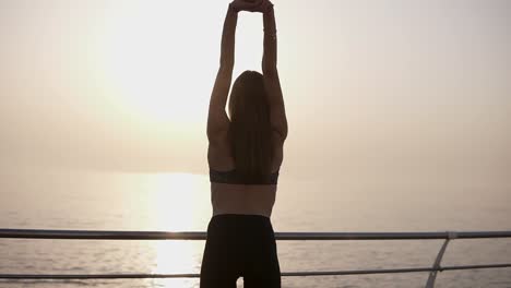 cheerful, young woman opens arms to sunrise at seaside. slim and graceful, in leggings. back side view