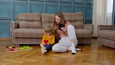 Woman-nanny-and-child-girl-studying-together-with-mobile-phone-and-tablet,-sitting-on-floor-at-home