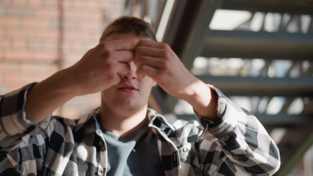young man in black white plaid shirt on outdoor metal staircase forcefully snaps cigarette in half between both hands with determined expression under sunlit brick wall and shadowed railing backdrop