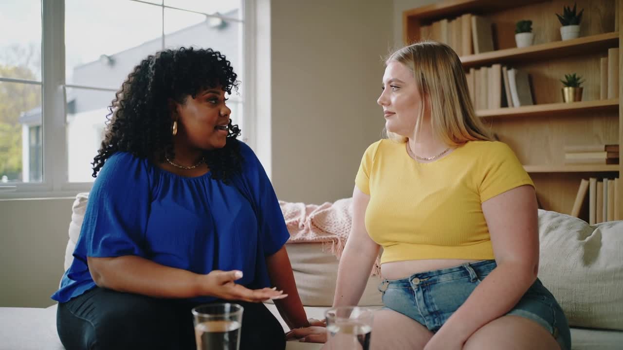 Talking left in blue blouse starting, gesturing while reassuring right in yellow crop top at home