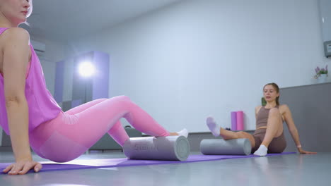 two women engaged in a fitness session on yoga mats using foam rollers for leg exercises in a well-lit gym, focus on stretching and muscle recovery