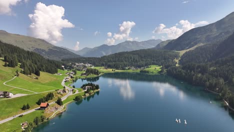 drone push in aerial view of lake surrounded by green mountain slopes and village under blue sky in davos, kanton graubünden, switzerland