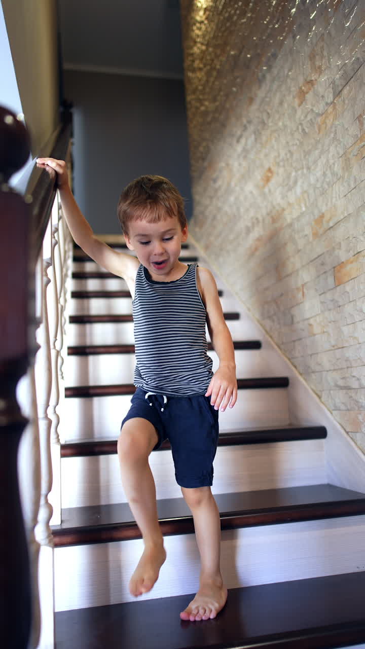 Adorable toddler kid going down the stairs holding by the handrail. Kid stops and sits on the stair. Vertical video.