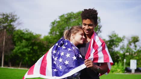 Attraktives-Multiethnisches-Paar,-Das-Sich-Umarmt-Und-Die-Amerikanische-Flagge-Auf-Dem-Rücken-Hält-Und-Auf-Der-Grünen-Wiese-Steht.-Patriotisch