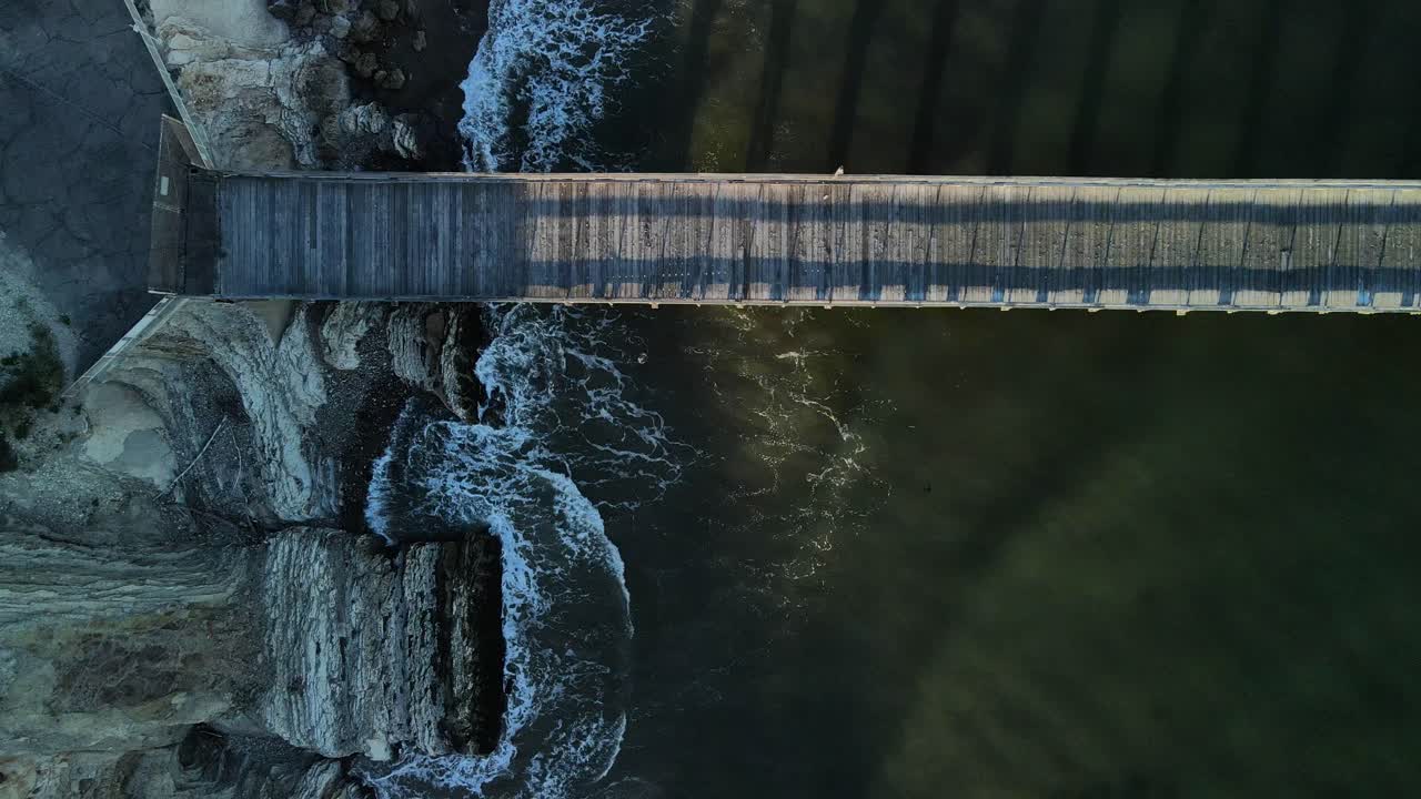 drone de arriba hacia abajo se eleva por encima del muelle de madera sin gente mientras las olas se estrellan en los acantilados grises de gaviota beach, california