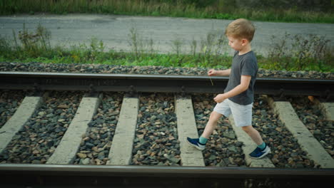 side view of young boy walking along rail track holding grass strand in hand, surrounded by gravel and greenery, with dirt road and plants in background under soft daylight in peaceful setting
