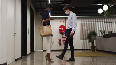 Diverse-business-people-wearing-face-masks-greeting-in-corridor-using-feet