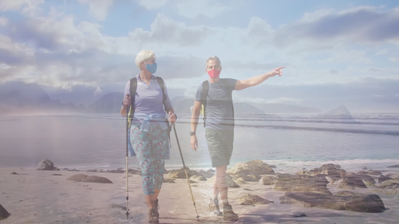 pareja de ancianos caucásicos con máscaras faciales caminando por la costa, sobre el mar en movimiento
