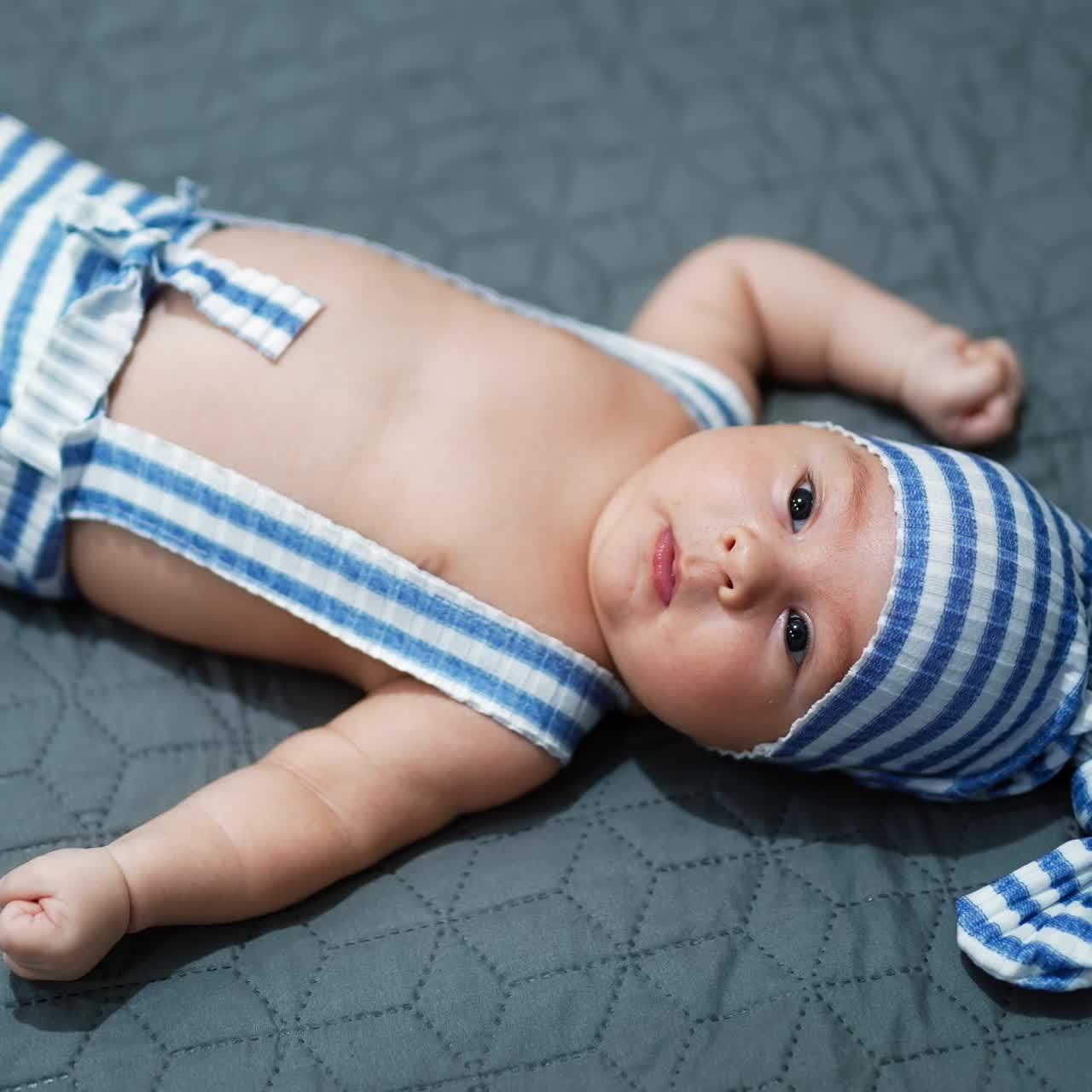 Baby boy wearing striped costume lying on the bed. Baby in a cap on the grey background. View from above