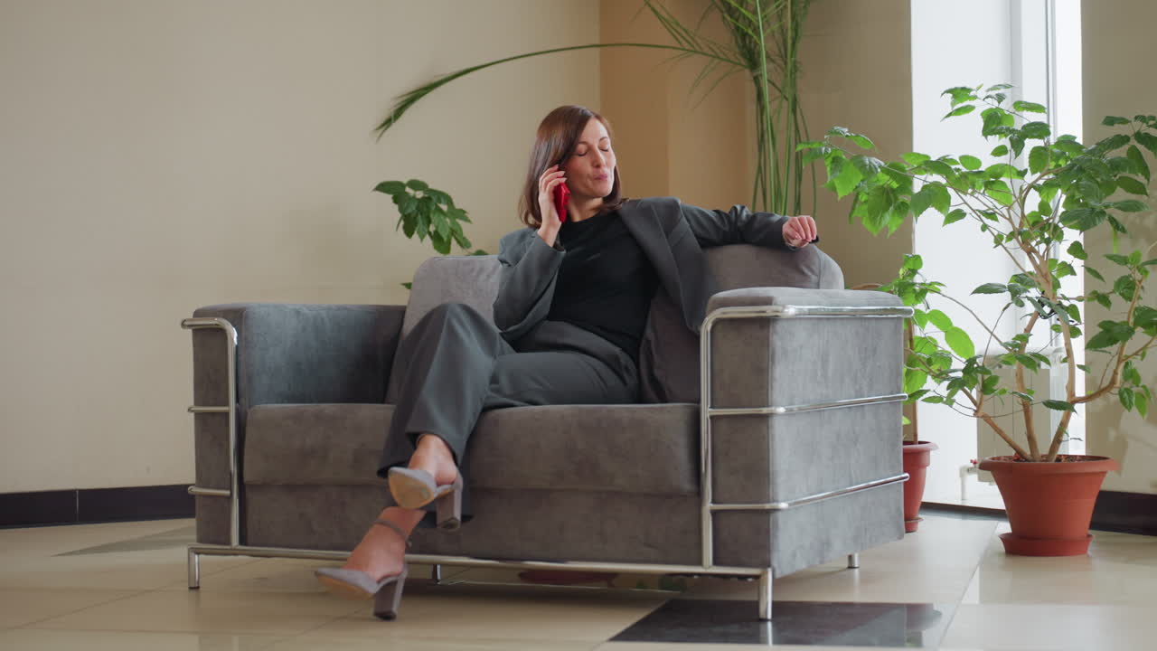 Smiling businesswoman in gray suit sits comfortably on modern couch talking on red smartphone in bright office space with large indoor plants, exuding confidence and ease during casual conversation