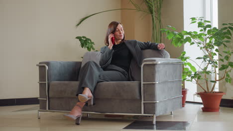 smiling businesswoman in gray suit sits comfortably on modern couch talking on red smartphone in bright office space with large indoor plants, exuding confidence and ease during casual conversation