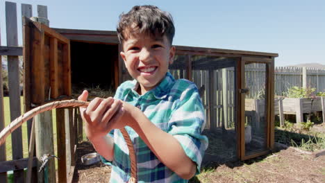 Portrait-of-happy-biracial-boy-holding-basket-with-eggs-next-to-henhouse,-slow-motion,-copy-space