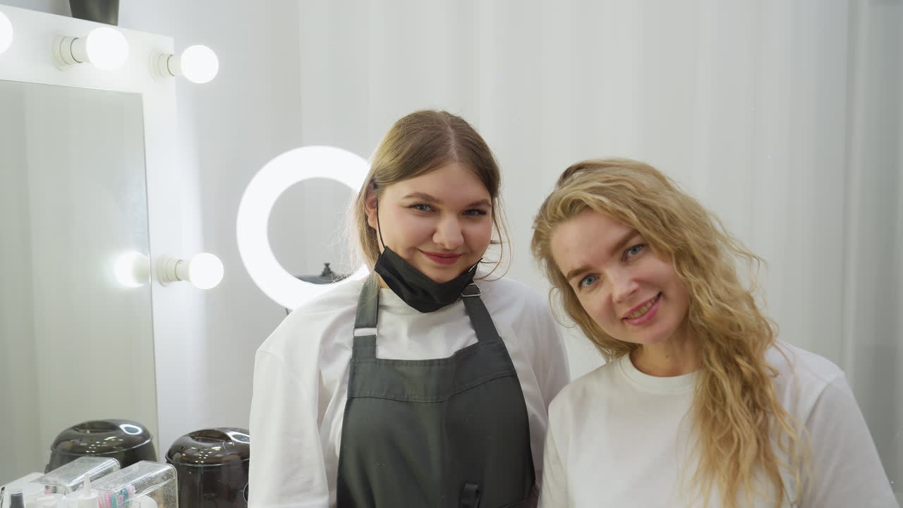 Client and beautician smile at camera inside salon with tools on table and clear reflection in mirror. Beautician wears black gloves and mask, client has wavy blonde hair and relaxed expression