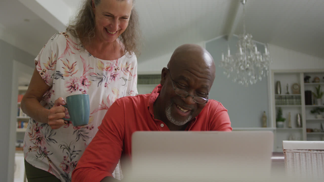Happy senior diverse couple wearing shirts and using laptop in living room