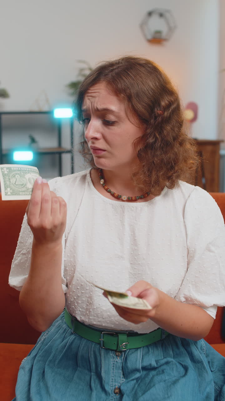 Upset young woman sitting on sofa counting money having not enough worried about finances debts