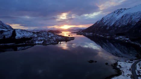 Toma-Aérea-De-La-Muñeca-Delantera-De-La-Puesta-De-Sol-En-Un-Día-Parcialmente-Nublado-Con-Majestuosas-Montañas-Cubiertas-De-Nieve-Y-Un-Hermoso-Reflejo-En-Las-Tranquilas-Aguas-De-Un-Lago-O-Mar