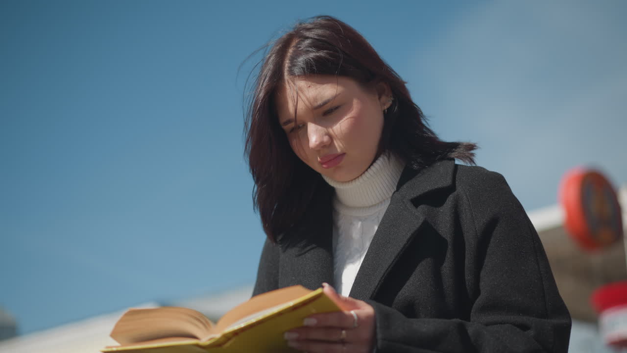 primer plano de una estudiante volviendo páginas de un libro al aire libre, con el viento revoloteando suavemente su cabello, detrás de ella hay un edificio de ladrillo rojo, capturando un momento pacífico de lectura bajo un cielo azul claro